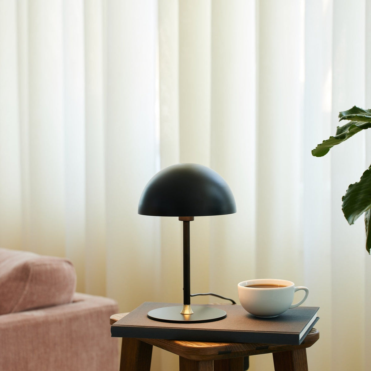Boogie Table Lamp 16cm in matt black with a dome shade and brass stem detail, styled on a side table beside a coffee cup in a living room setting.