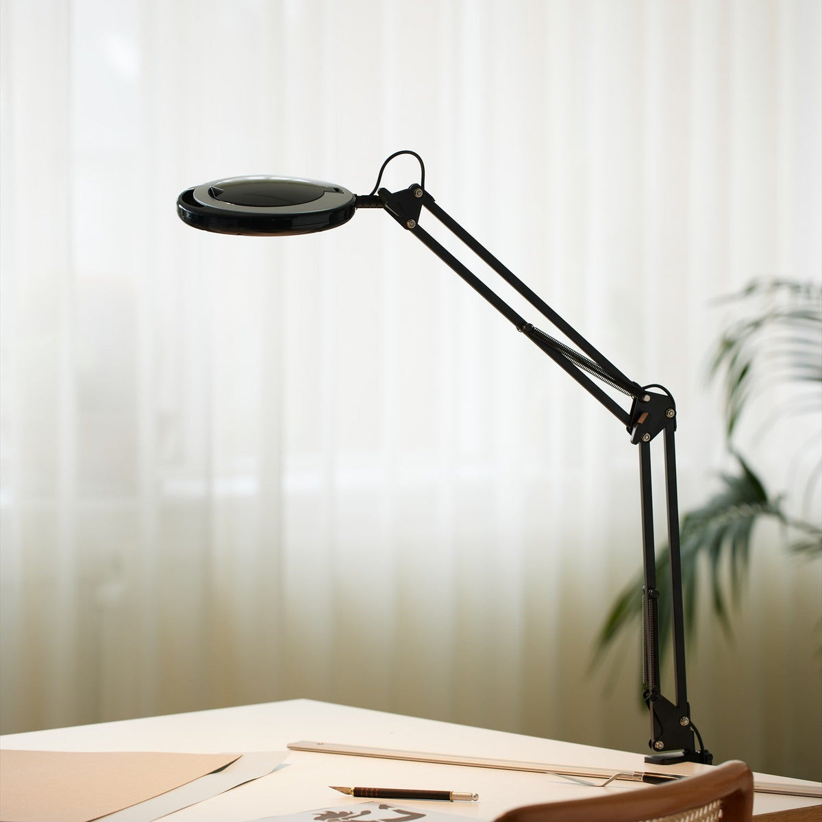 Magni LED Archi Clip Desk Table Lamp in black, with round magnifying head and folding sprung arm, clamped to a desk beside paper and pen, softly lit interior background.
