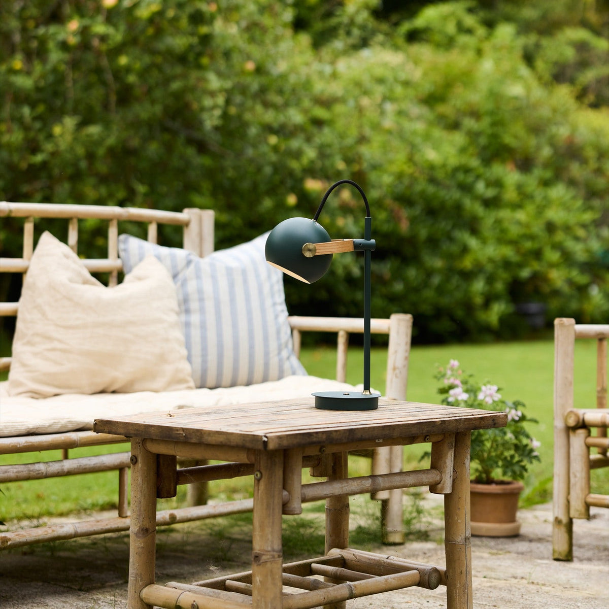 DC LED Go Portable Table Lamp in dark green, with round tilting shade on an oak strap and slim post, shown on a wooden table beside pillows in a garden seating area.