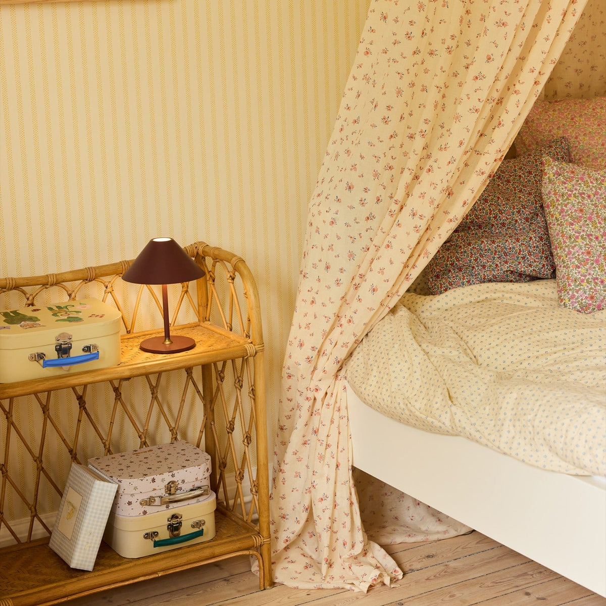 Hygge LED Rechargeable Outdoor Table Lamp in maroon with a wide cone shade and slim stem on a round base, shown on a wicker shelf in a bedroom setting.