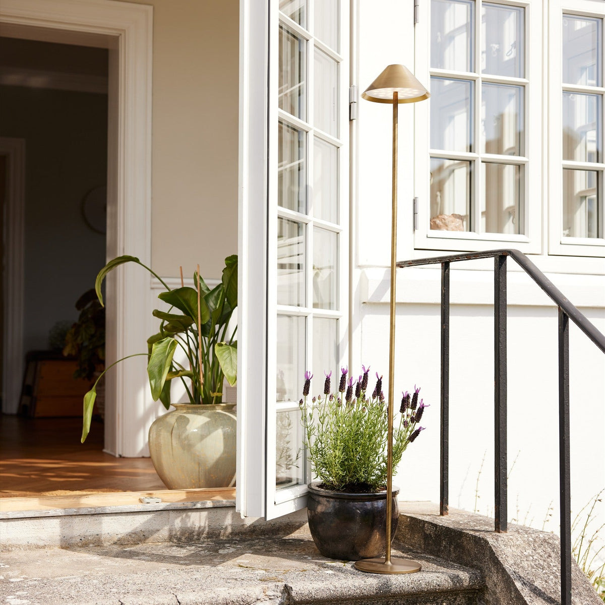Hygge LED rechargeable outdoor floor lamp in antique brass with slim stem and cone shade, shown by a doorway with lavender and a potted plant.