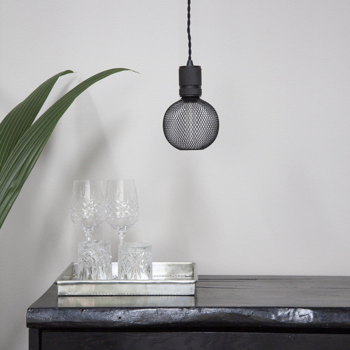Colors Gitter Bulb 125mm in black wire mesh, hanging from a matte black pendant fitting above a dark console table with crystal glassware, set against a light grey wall.