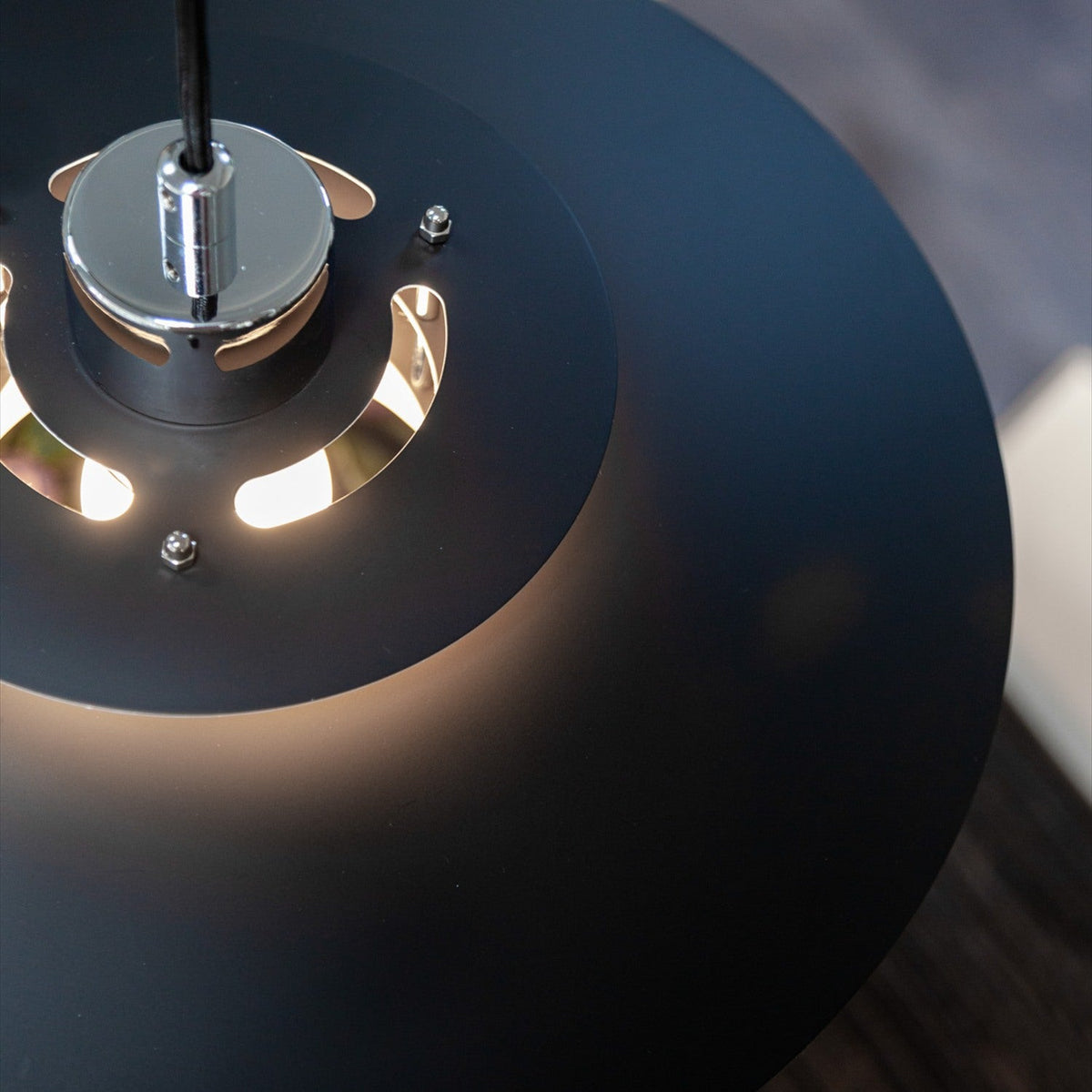 Close-up of the Sørup Pendant Light 40cm in matte black metal, showing the chrome mounting cylinder and cut-out vents that reveal glowing bulb light beneath the shade.