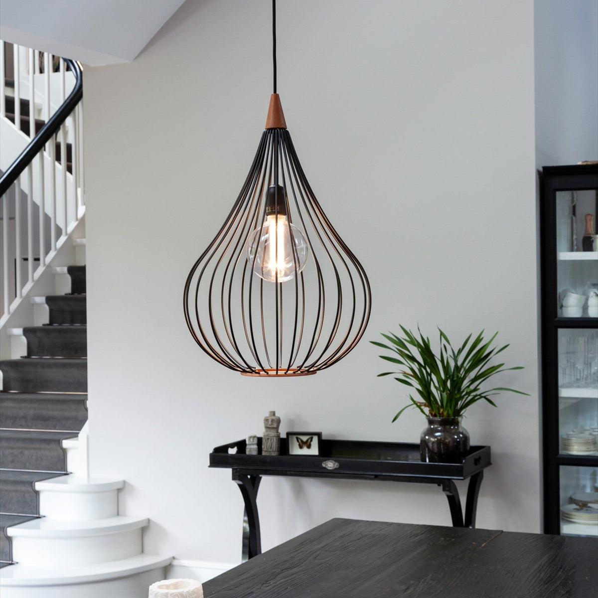 Drops Pendant Light 38cm in black and dark wood, a teardrop-shaped open wire cage with exposed bulb, hung above a dark table near a staircase and dining area.