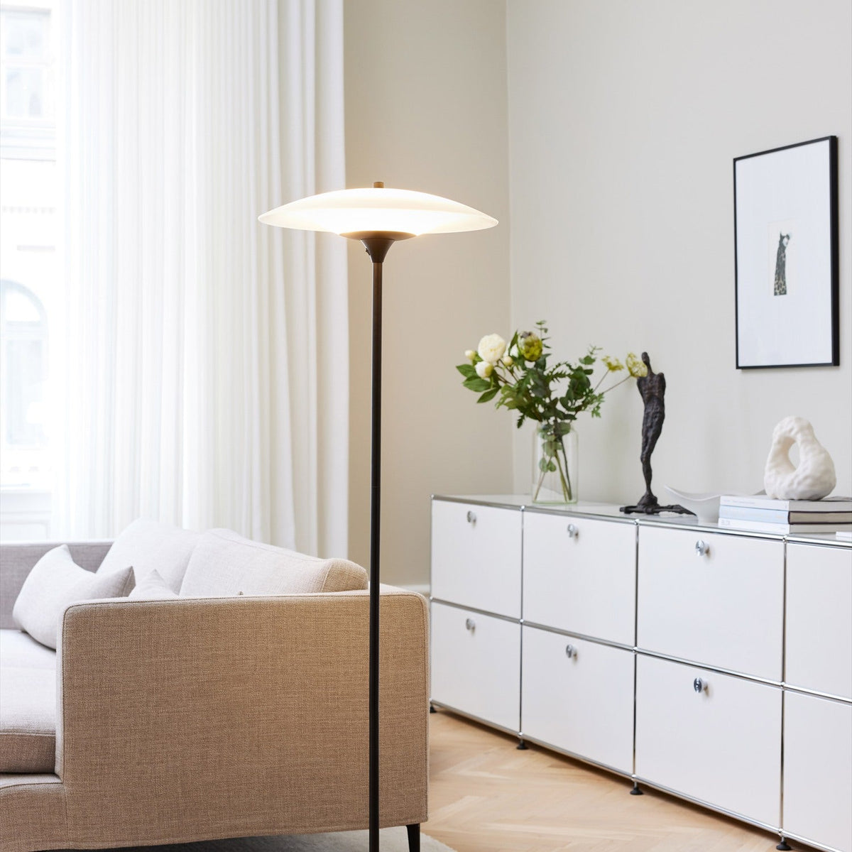 Baroni Glas Floor Lamp 46cm in opal and black, with a slim black stem and round disc base, shown lit beside a sofa and sideboard in a living room.