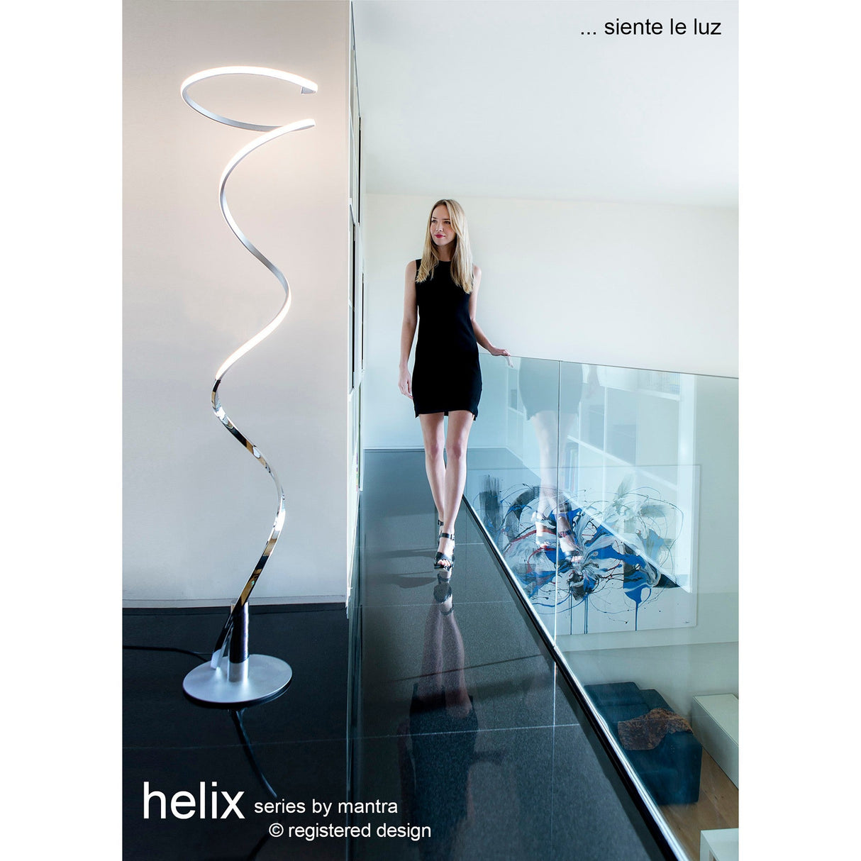 A woman in a black dress walks on a glass walkway beside the Helix LED Floor Lamp 42W 3000K in Silver & Chrome. The minimal, bright space features art below. Text reads “helix series by mantra” and “siente le luz.”.