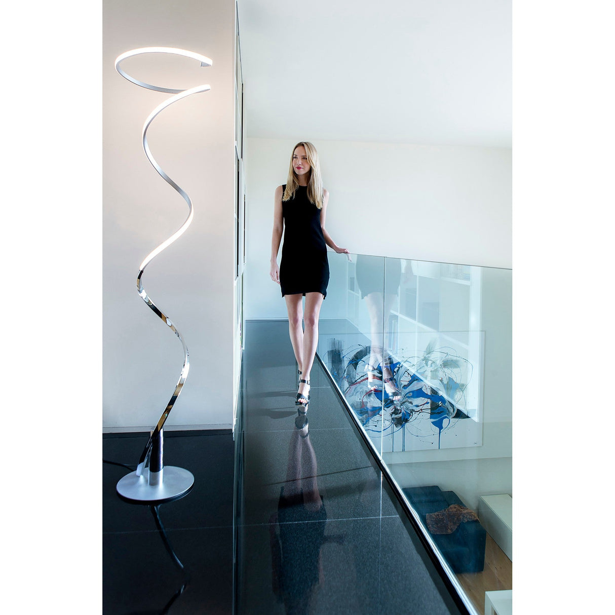 A woman in a black dress walks through a bright, minimalistic hallway with a glass railing. Nearby stands the curvy Helix LED Floor Lamp 42W 3000K in Antique Brass, illuminating the modern space.