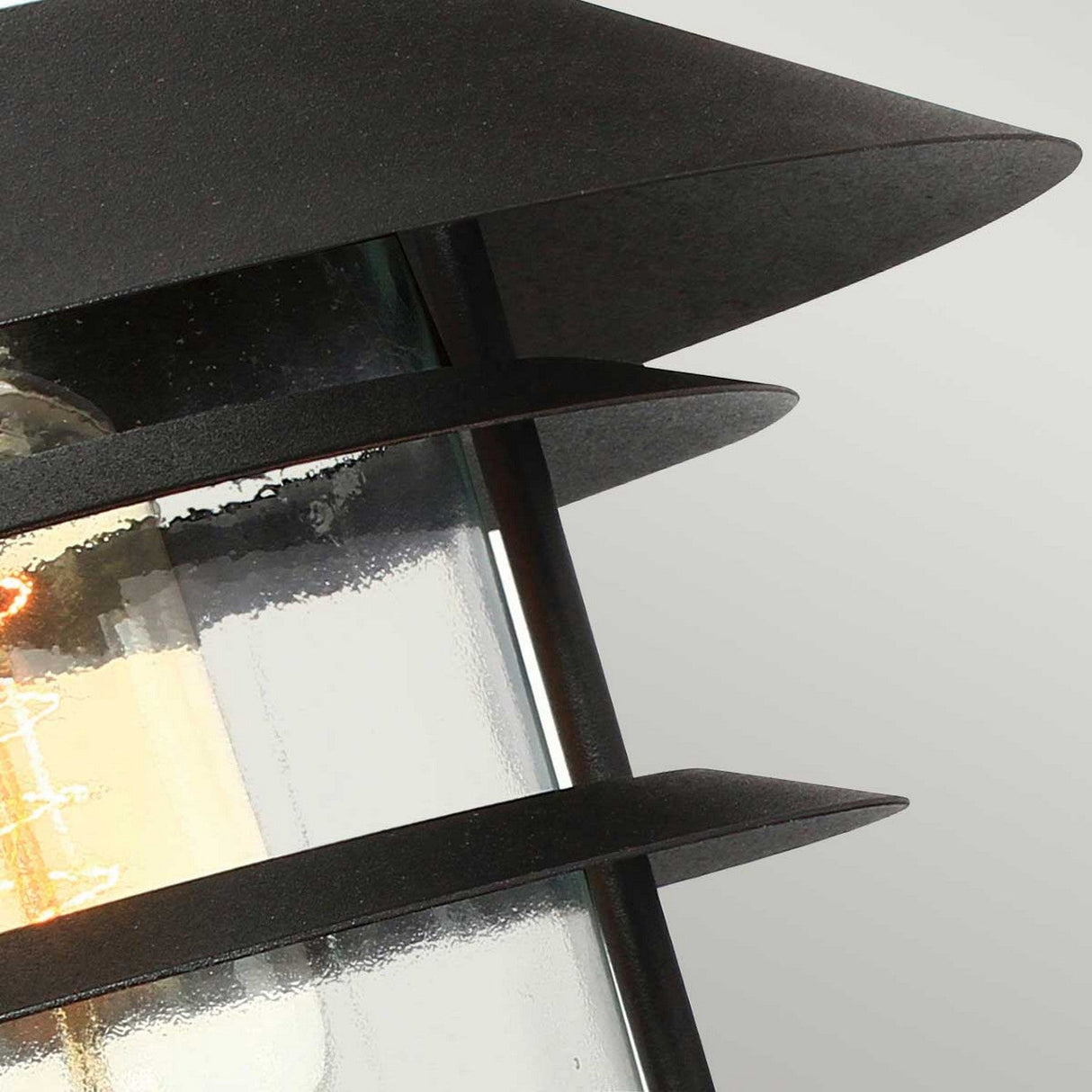 Close-up of the Helsingor 1 Light Outdoor Wall Light, featuring a Danish-inspired design with glass and black metal accented by horizontal slats. The partially visible bulb casts a soft glow, enhancing its elegance. With an IP44 rating, it's ideal for any outdoor setting. The background is a light gray.