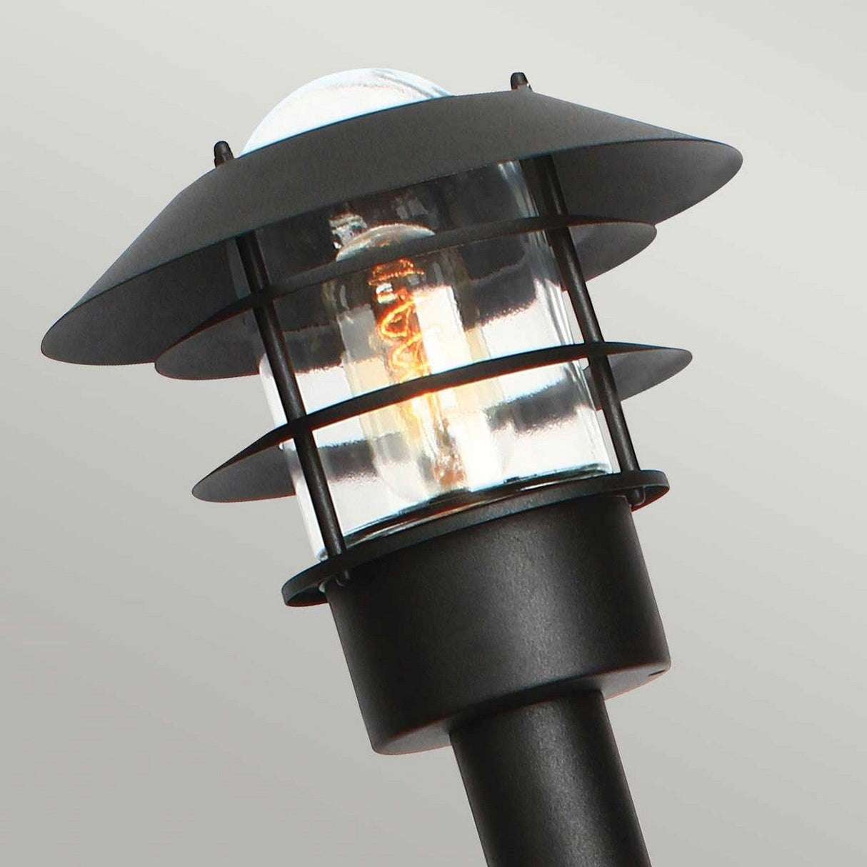 A close-up of the Helsingor 1 Light Bollard Lantern in black showcases its clear glass bulb, accentuated by multiple horizontal metal rings surrounding it. This lantern emits a soft orange glow, making it ideal for coastal environments, set against a gray background.