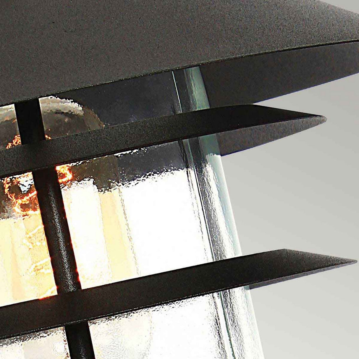 A close-up of the Helsingor 1 Light Pedestal Light - Black showcases its Scandinavian design, featuring a glowing bulb encased within a black fixture with horizontal slats and a glass enclosure. This combination provides a sleek, modern aesthetic that epitomizes sophisticated outdoor lighting against a neutral backdrop.