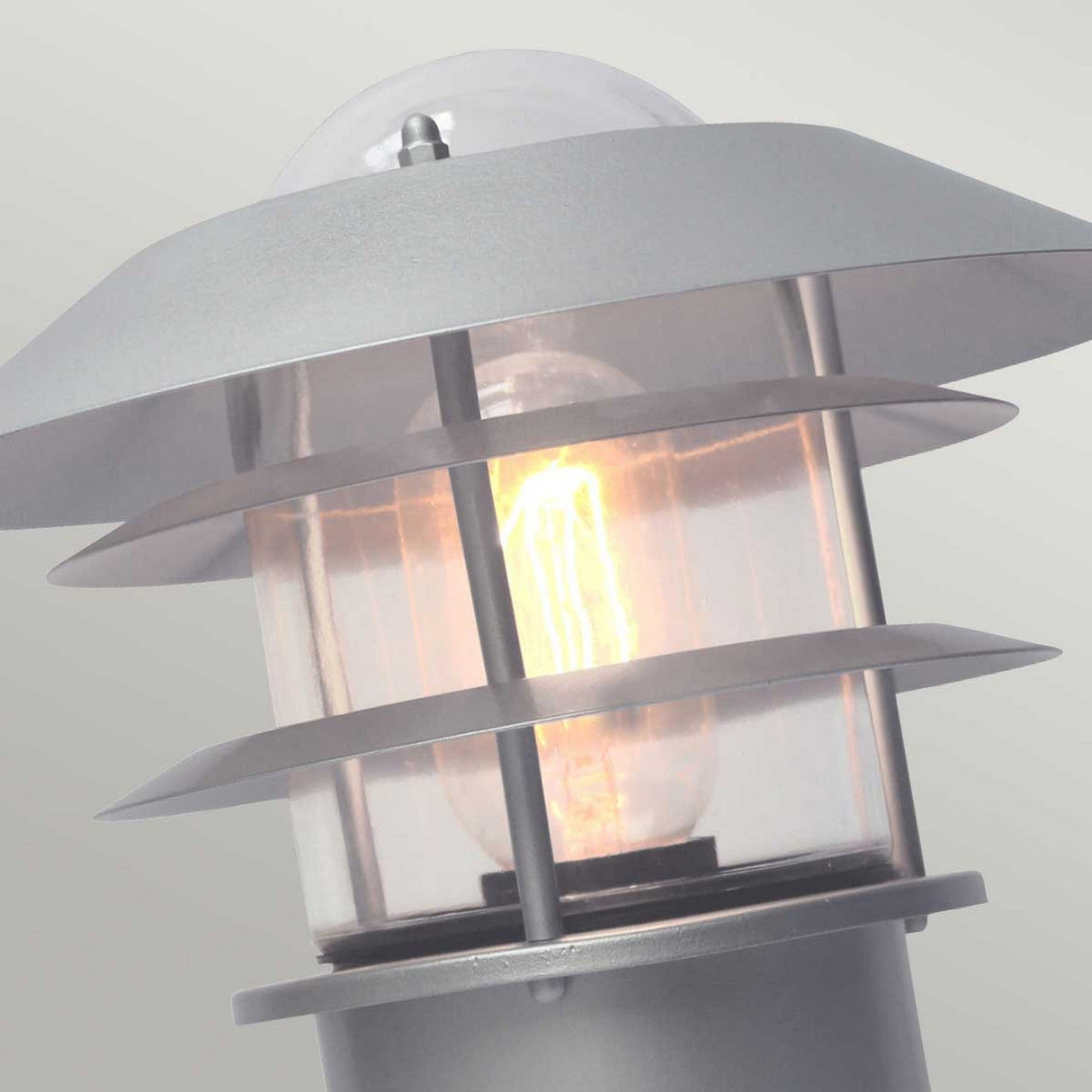 The Helsingor 1 Light Pedestal Light - Silver is a contemporary outdoor fixture showcasing a radiant bulb within cylindrical glass. Its sleek design includes stainless steel with layered horizontal panels and offers soft illumination against a light gray background, boasting an IP44 rating.