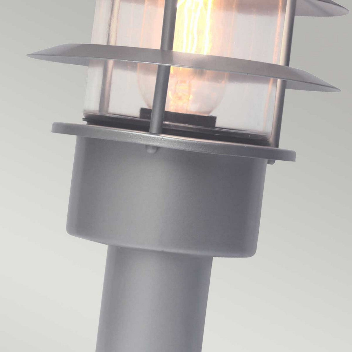 Close-up of the Helsingor 1 Light Pedestal Light - Silver, showcasing its cylindrical base and glass encasement with a glowing bulb. The fixture features a stainless steel finish accented by horizontal metal rings around the glass. It is designed with an IP44 rating for durability, set against a soft gradient background.