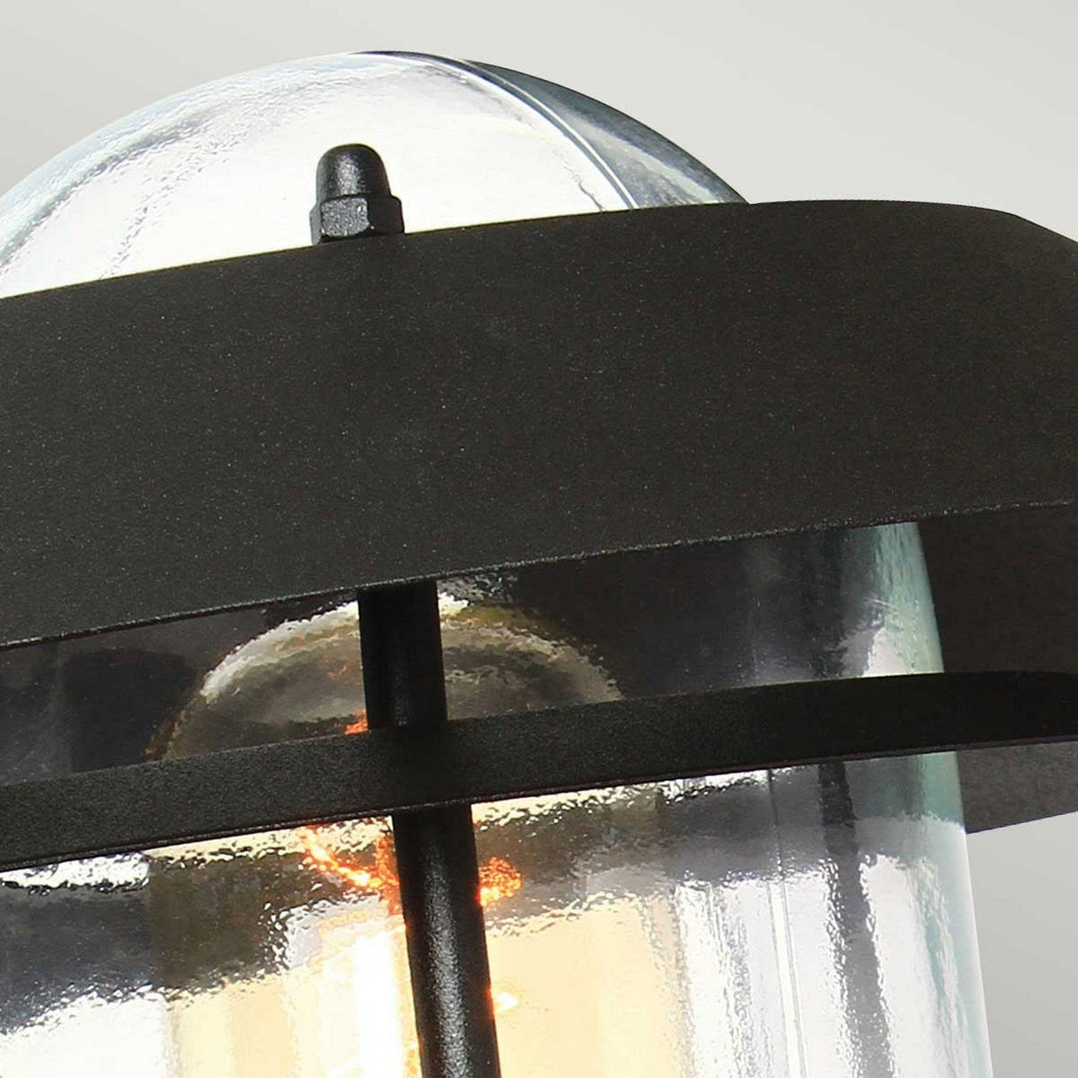 A close-up of the Helsingor 1 Light Outdoor PIR Wall Light - Black reveals a sleek design with a cylindrical glass shade and black metal bands. This weather-resistant exterior fixture includes a glowing bulb and a Passive Infrared sensor, providing not only warm ambiance but also enhanced security by automatically detecting movement.