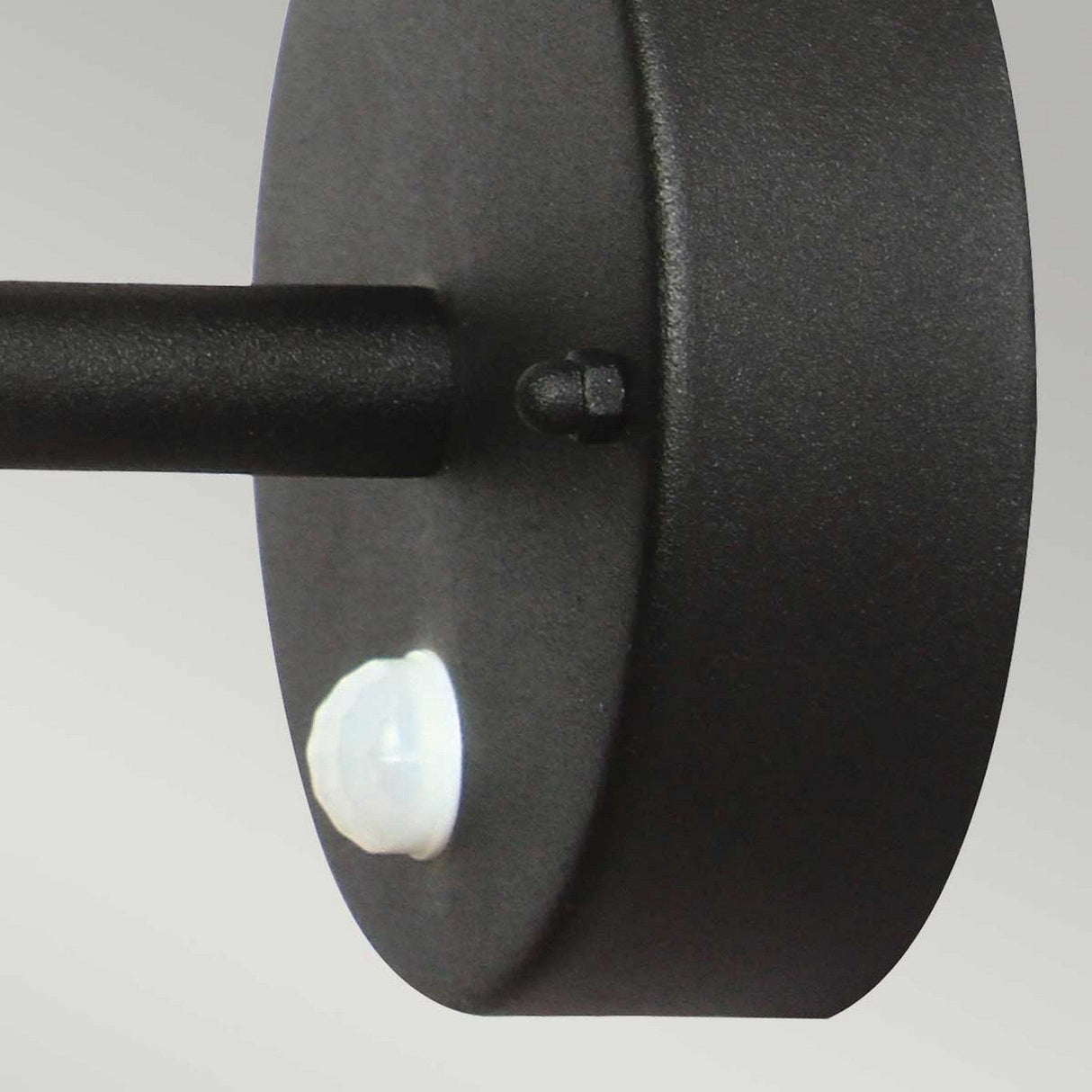 A close-up of the Helsingor 1 Light Outdoor PIR Wall Light - Black shows its sleek black design with a round base. A white motion sensor is evident on the bottom of this weather-resistant exterior lighting fixture, set against a plain and light-colored background.