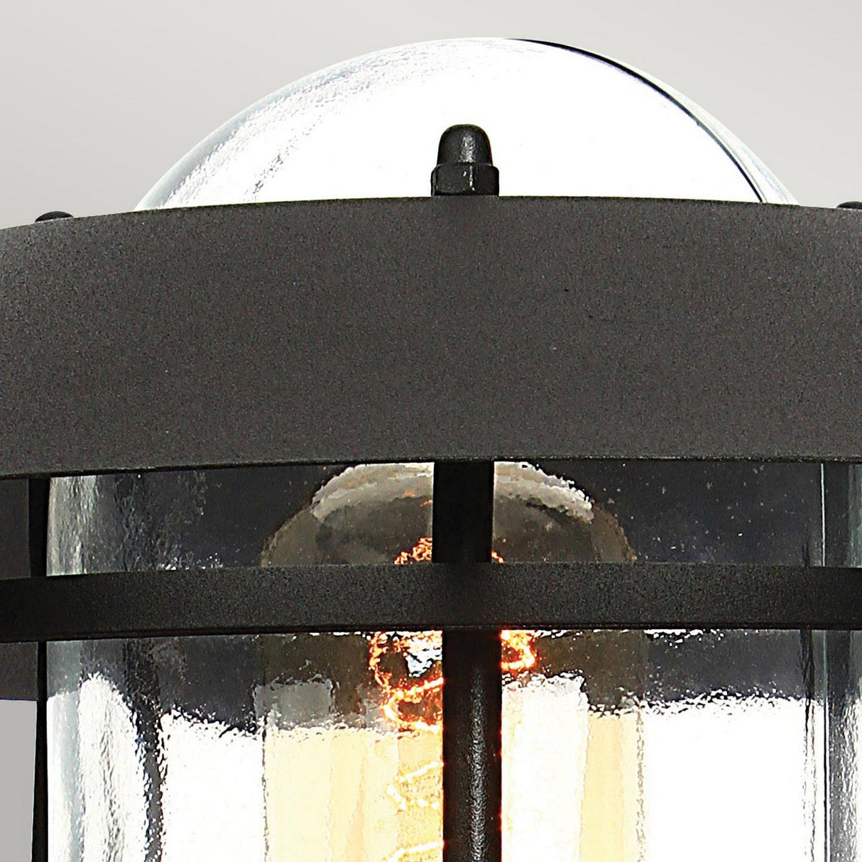 Close-up of the Helsingor 1 Light PIR Pedestal Light - Black, showcasing its clear glass enclosure and black metal frame with a glowing bulb inside. This sophisticated outdoor light enhances security in your space, set against a light gray background.