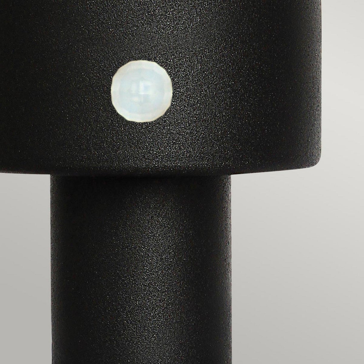 Close-up of the Helsingor 1 Light PIR Pedestal Light in black, showcasing a small circular white PIR sensor embedded on its surface. The plain light gray backdrop accentuates the security lighting design.