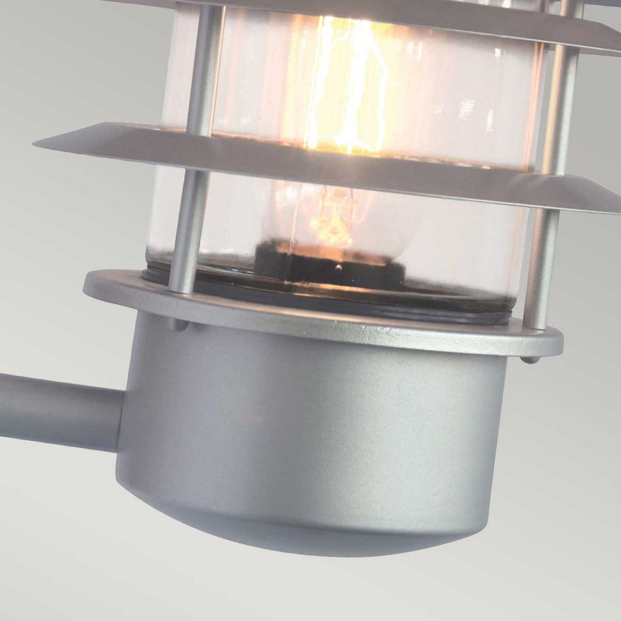 Close-up of the Helsingor 1 Light Outdoor PIR Wall Light in silver. This energy-efficient outdoor lamp includes a cylindrical glass cover that encases a lit bulb, accentuated by horizontal metal rings. The background is a neutral, blurred surface, highlighting its sleek design and functionality for security lighting.