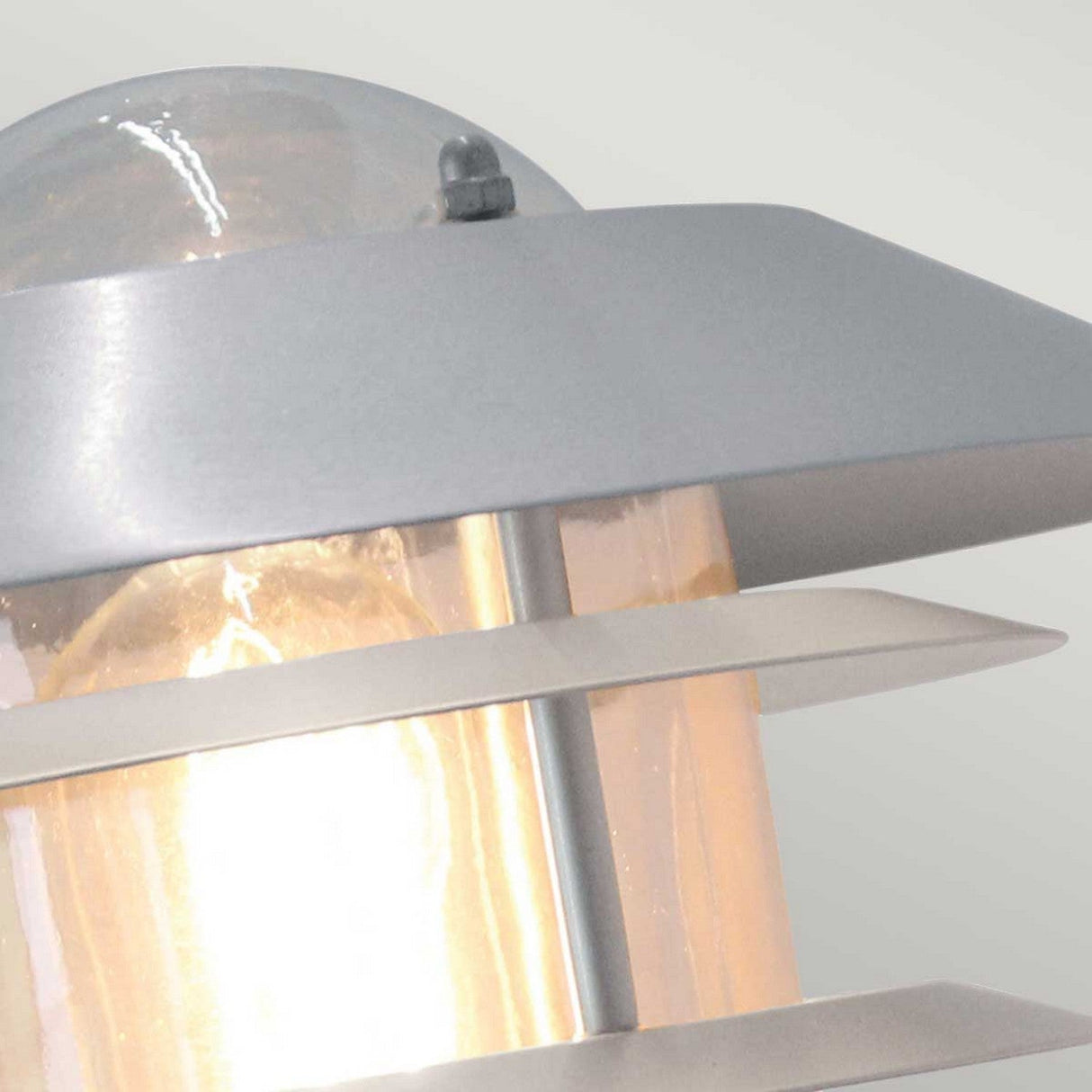 Close-up of the Helsingor 1 Light Outdoor Wall Light in Silver, featuring a sleek stainless steel frame and glass panels. The visible bulb emits a warm glow, enhancing its IP44-rated design. This fixture boasts a modern, contemporary style that's perfect for any setting.