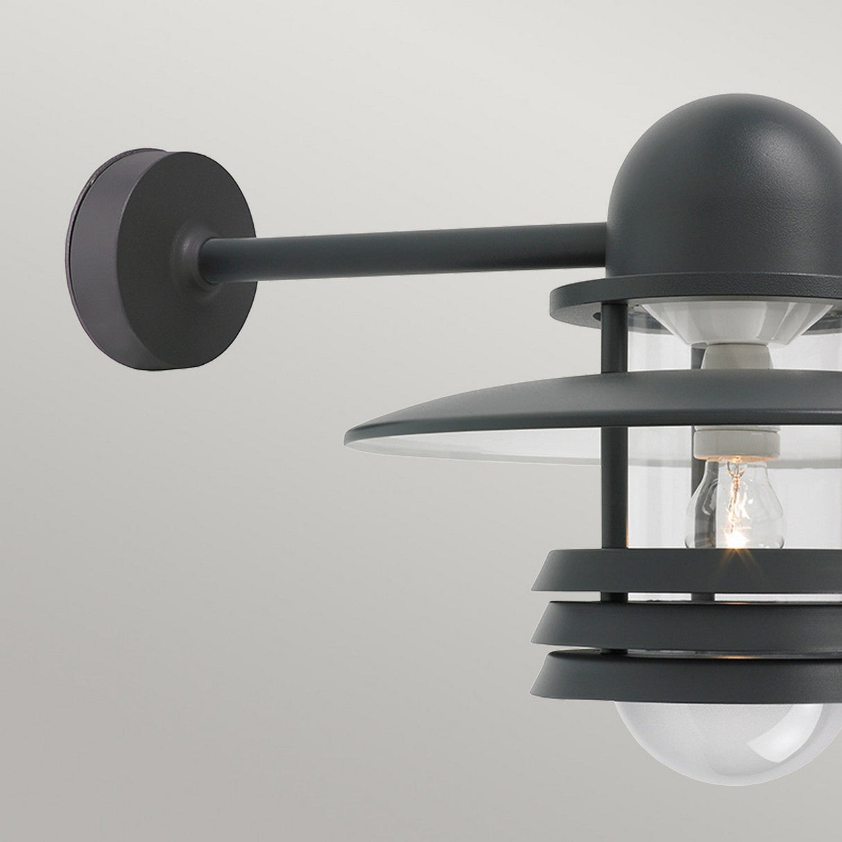 Close-up of the Helsinki Outdoor 1 Light Wall Light, a contemporary exterior lighting fixture with Scandinavian flair. It includes a dome-shaped cover and circular metal frame, finished in graphite with a translucent glass panel that showcases a visible bulb inside.