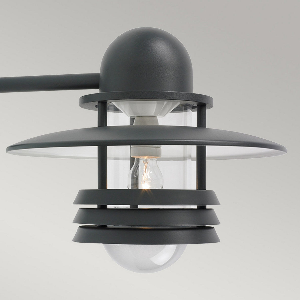 Close-up of the Helsinki Outdoor 1 Light Wall Light in Graphite, a modern outdoor lighting fixture. This Scandinavian design showcases a bulb enclosed in transparent glass, encircled by concentric circular metal elements for a stylish and contemporary look.