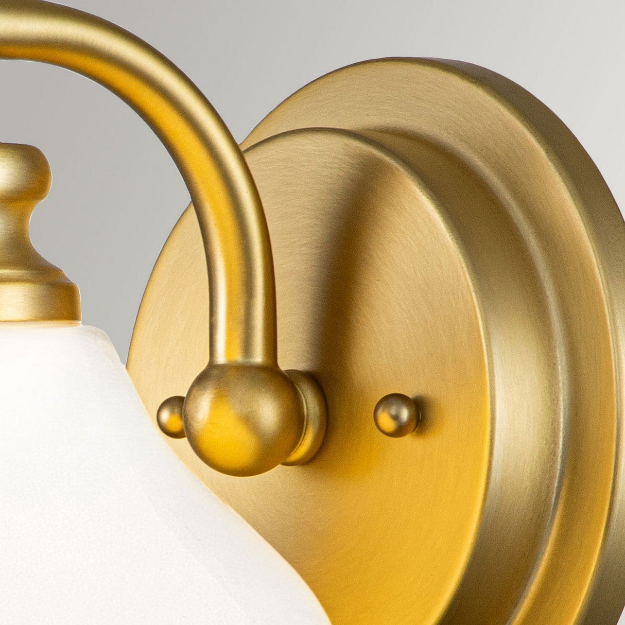 A close-up of the Ainsley 1 Light Wall Light in brushed brass showcases its round base and curved arm, which supports an opal glass shade. The metal surface has a smooth, polished finish that adds elegance and warmth to the fixture, complemented by an energy-efficient LED lamp.