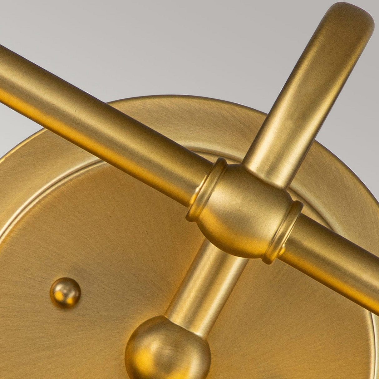 A close-up view of the Ainsley 2 Light Wall Light in brushed brass shows its metallic, gold-colored fixture featuring intersecting cylindrical rods and circular elements. The smooth finish combines minimalist industrial aesthetics with opal glass shades against a pale background.