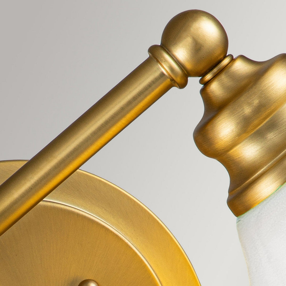 Detailed view of the Ainsley 2 Light Wall Light's brushed brass arm and fixture, showcasing its graceful curves and matte finish. The partly visible opal glass shade provides a striking contrast against the brass, all set against a light gray backdrop.