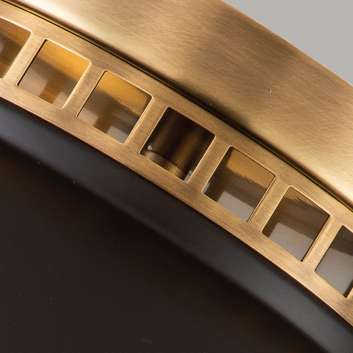 Close-up of the Amelia 2 Light Flush Light - Bronze, a circular object featuring a brass and black design with rectangular cut-outs along its edge, unveiling a shiny interior. Its bronze finish enhances the modern, industrial vibe, while the cast glass diffuser provides an elegant touch.