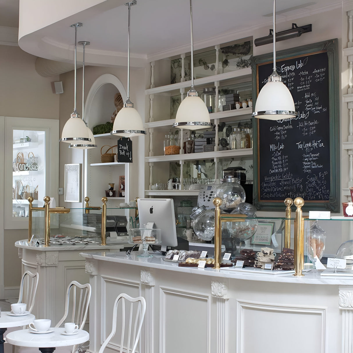 A cozy café interior with a white decor includes a counter adorned with pastries, an iMac, and a menu displayed on a chalkboard. Hanging from the ceiling is the Amelia 1 Light Medium Pendant in chrome, accompanied by white chairs and a vintage cash register.