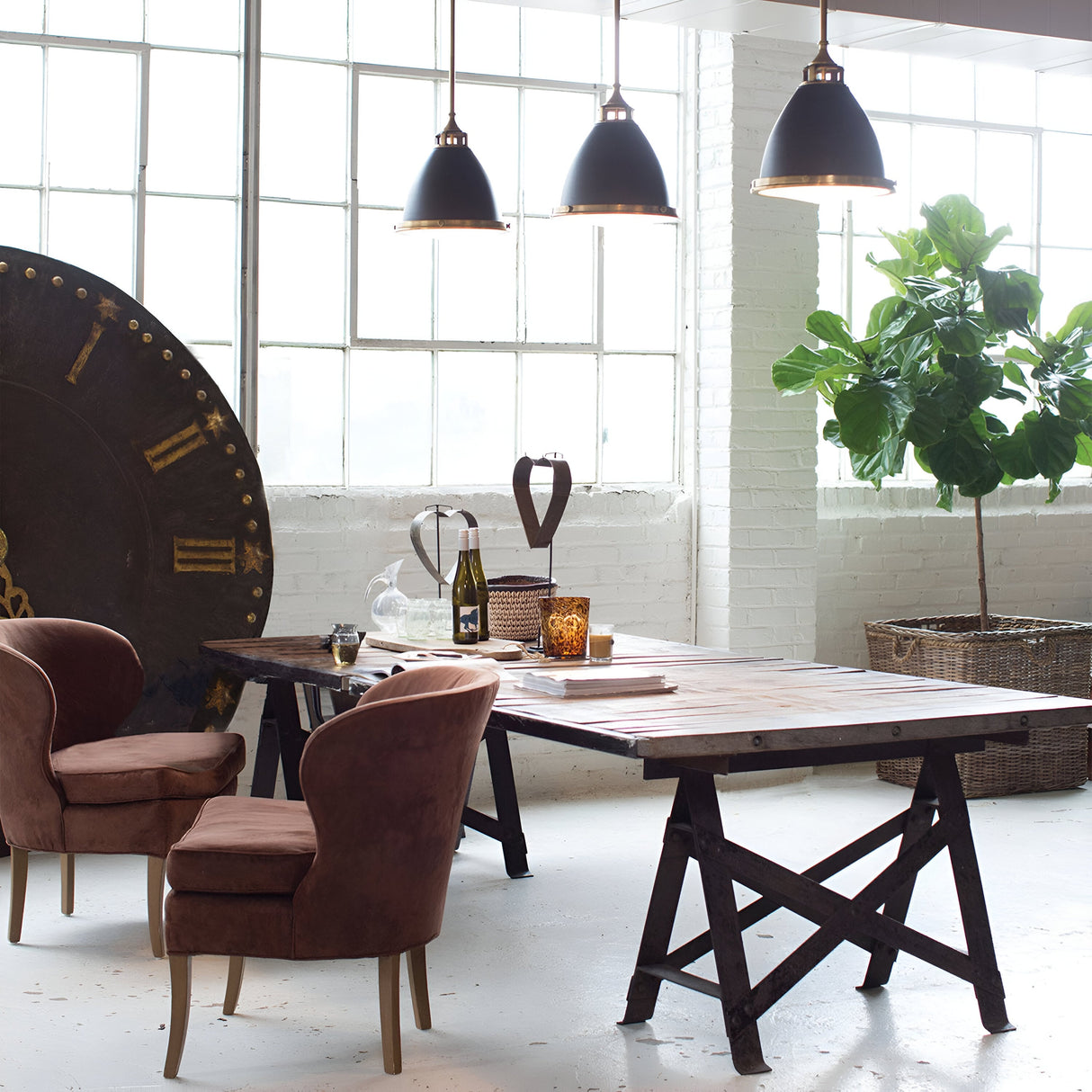 A cozy industrial-style room with large windows features a wooden table surrounded by brown upholstered chairs. A decorative clock face leans against a wall, and a potted plant adds greenery. Hanging above the table is the Amelia 1 Light Medium Pendant in bronze, with opal glass domes that create a warm ambiance.