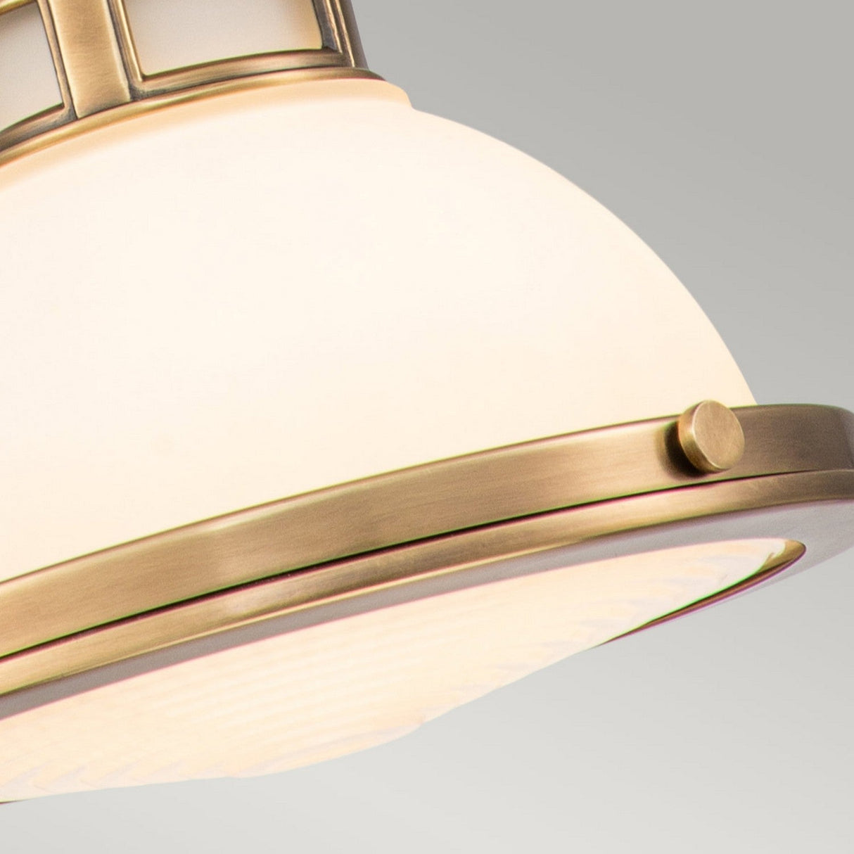 Close-up of the Amelia 1 Light Mini Pendant in brass, featuring an opal glass dome with a frosted cover. Its design includes a rounded metal frame adorned with a minimalistic geometric pattern on top, set against a soft gray background.