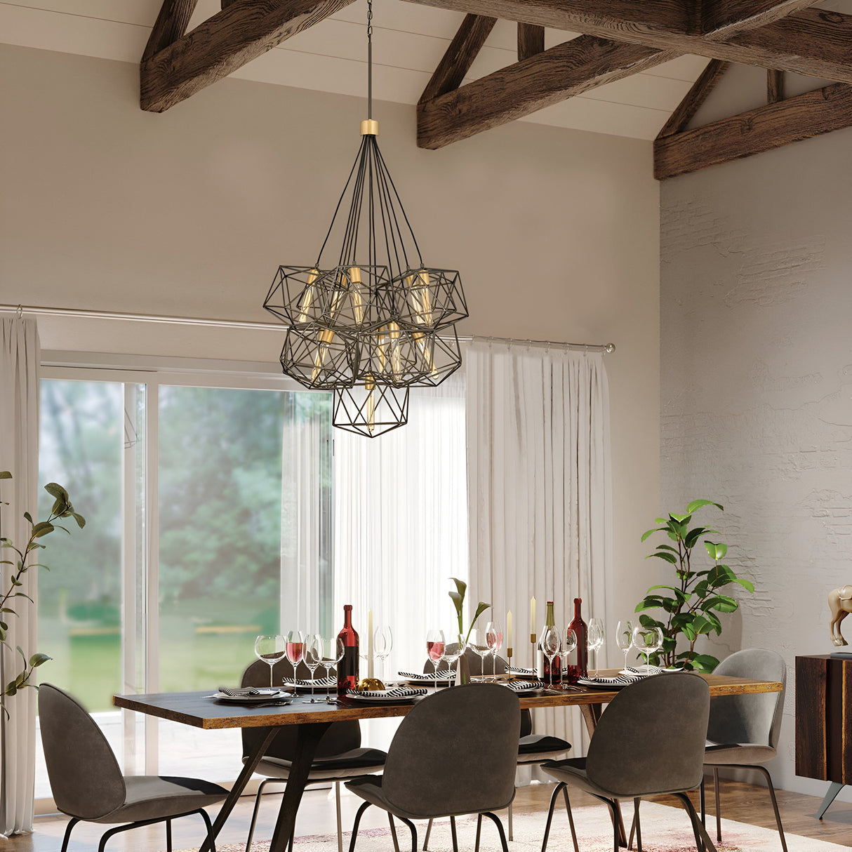 A modern dining room features a wooden table set for six, accompanied by gray chairs and illuminated by the Astrid 11 Light Cluster Pendant in Matte Bronze & Gold, casting a warm glow. Large windows with sheer curtains frame the lush greenery outside, while wine bottles and glassware elegantly adorn the table as décor.
