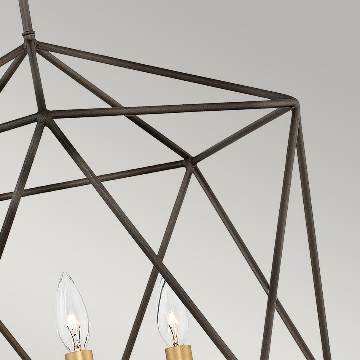 A detailed view of the Astrid 3 Light Chandelier in matte bronze and gold highlights its cage-like structure. This pendant chandelier, with candle-shaped light bulbs set against a neutral gray background, epitomizes a modern, minimalist design featuring sharp angles and an industrial aesthetic.