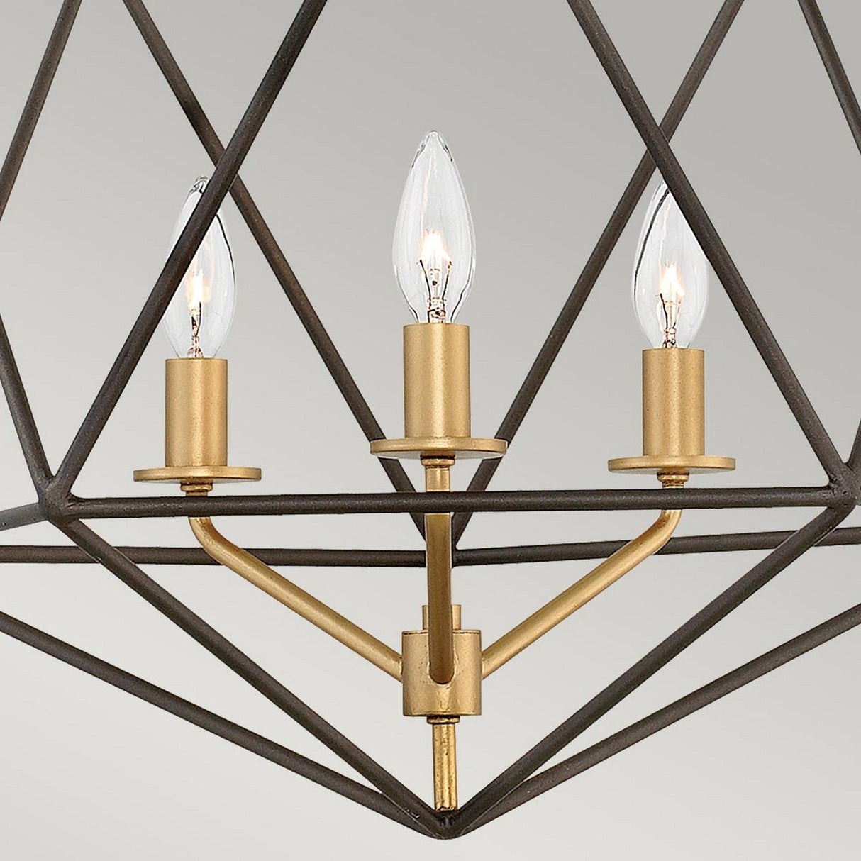 Introducing the Astrid 3 Light Chandelier - Matte Bronze & Gold: This geometric pendant chandelier showcases three candle-shaped bulbs encased in a matte bronze frame with a gold finish. The design highlights triangular patterns and modern lines against a light gray backdrop, radiating contemporary elegance.