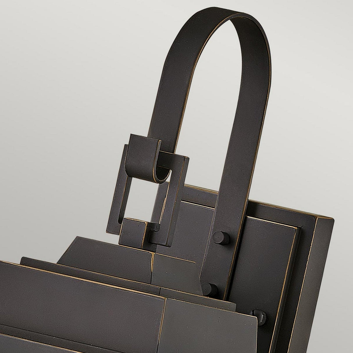 A close-up of the Bainbridge 1 Light Outdoor Medium Wall Lantern in bronze showcases its dark, modern metal design with bevelled glass panels and angular elements, complemented by a curved handle, all set against a light gray background.