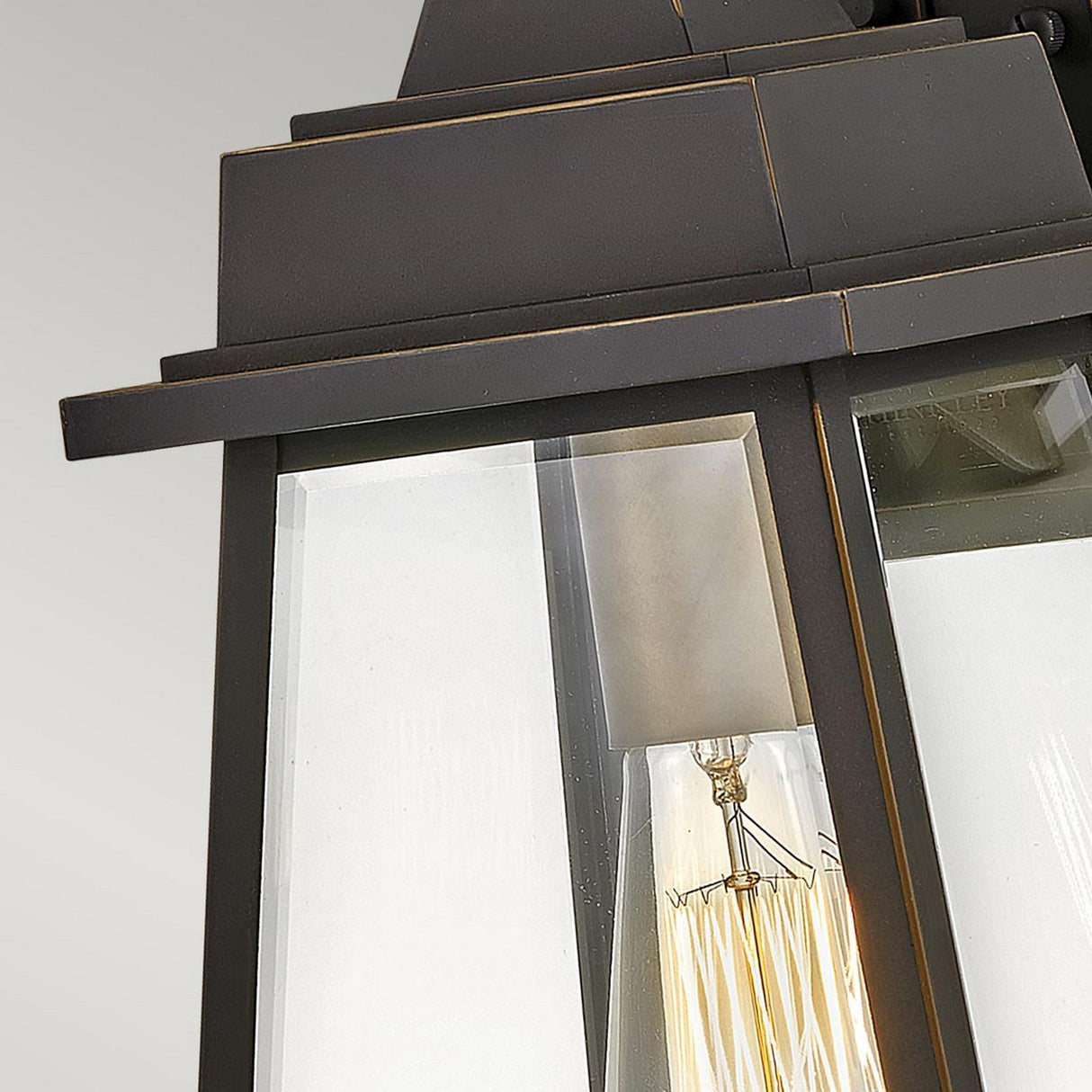A close-up of the Bainbridge 1 Light Outdoor Medium Wall Lantern in bronze, showcasing bevelled glass panels and a vintage-style exposed filament bulb inside against a plain light gray background.