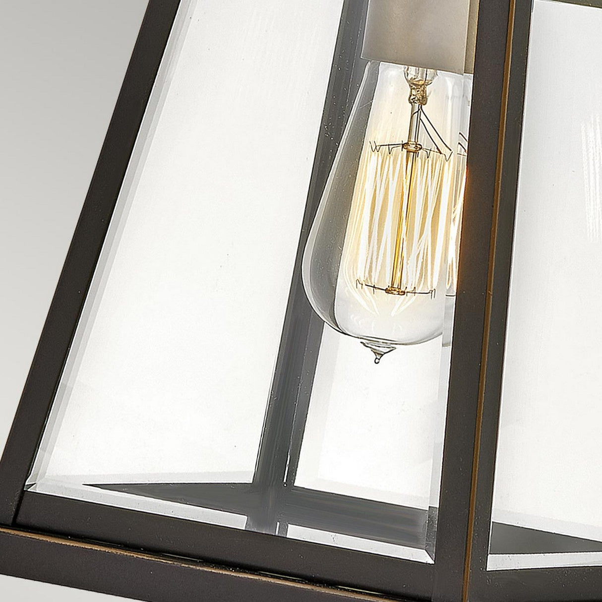A close-up of the Bainbridge 1 Light Outdoor Medium Wall Lantern showcases its bevelled glass panels and provides a clear view of the vintage Edison bulb inside. The rectangular silhouette is outlined by a black metal frame, with the bulb’s filament offering a decorative accent enhanced by its elegant bronze finish.
