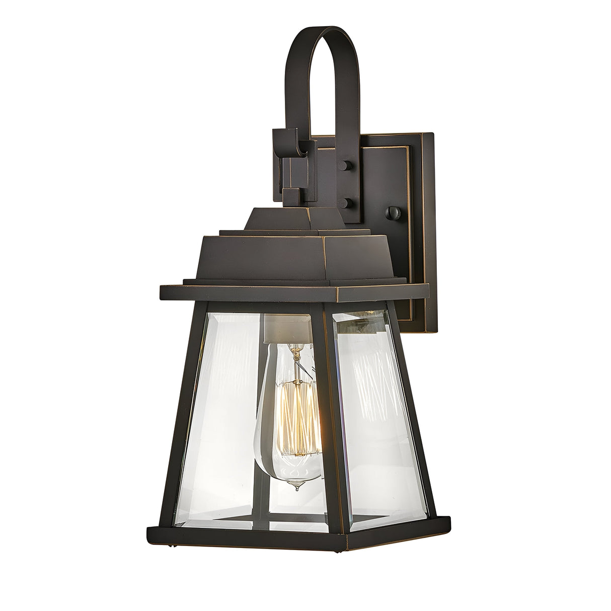 The Bainbridge 1 Light Outdoor Small Wall Lantern in bronze features a black metal frame with bevelled glass panels encasing a transparent enclosure. Within, an exposed filament bulb casts a warm glow. Its design seamlessly blends traditional and modern styles, making it perfect for use both outdoors and indoors.