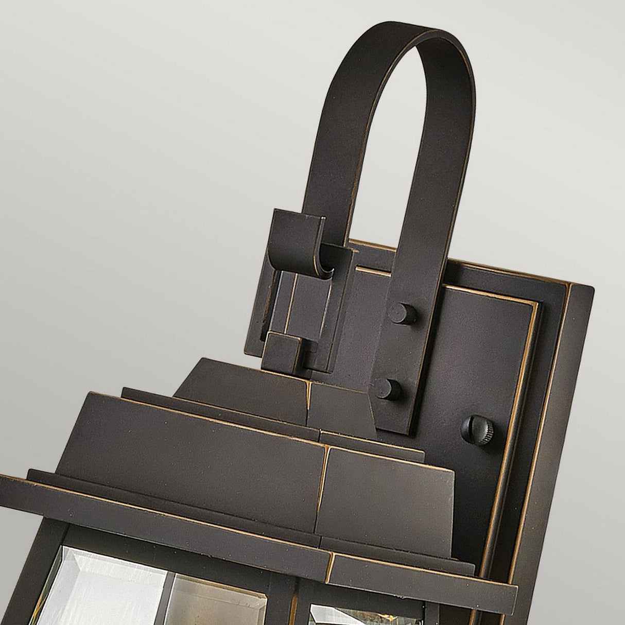 Close-up of the Bainbridge 1 Light Outdoor Small Wall Lantern in bronze, featuring a square base and curved arm. The design highlights bevelled glass panels with a dark finish complemented by subtle bronze accents, set against a light gray background.