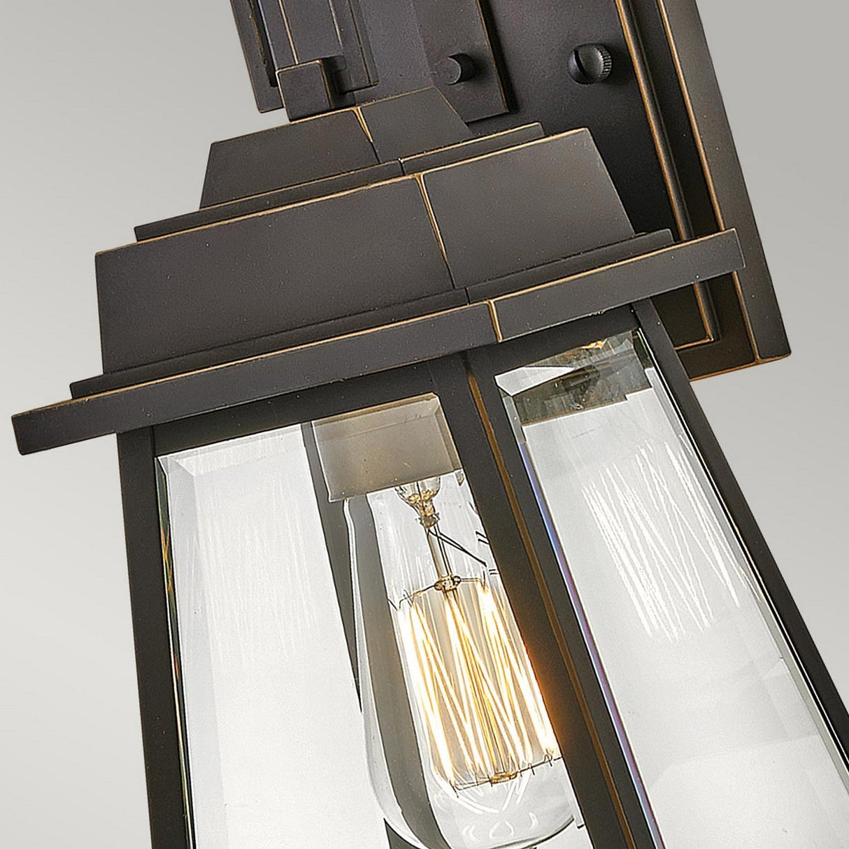 A close-up of the Bainbridge 1 Light Outdoor Small Wall Lantern - Bronze showcases its modern black metal and glass structure, featuring a visible Edison bulb inside. The design includes sleek lines that evoke the aesthetic of a bronze lantern, with the bulb encased in clear glass. Its geometric shape stands out strikingly against the plain light gray background.