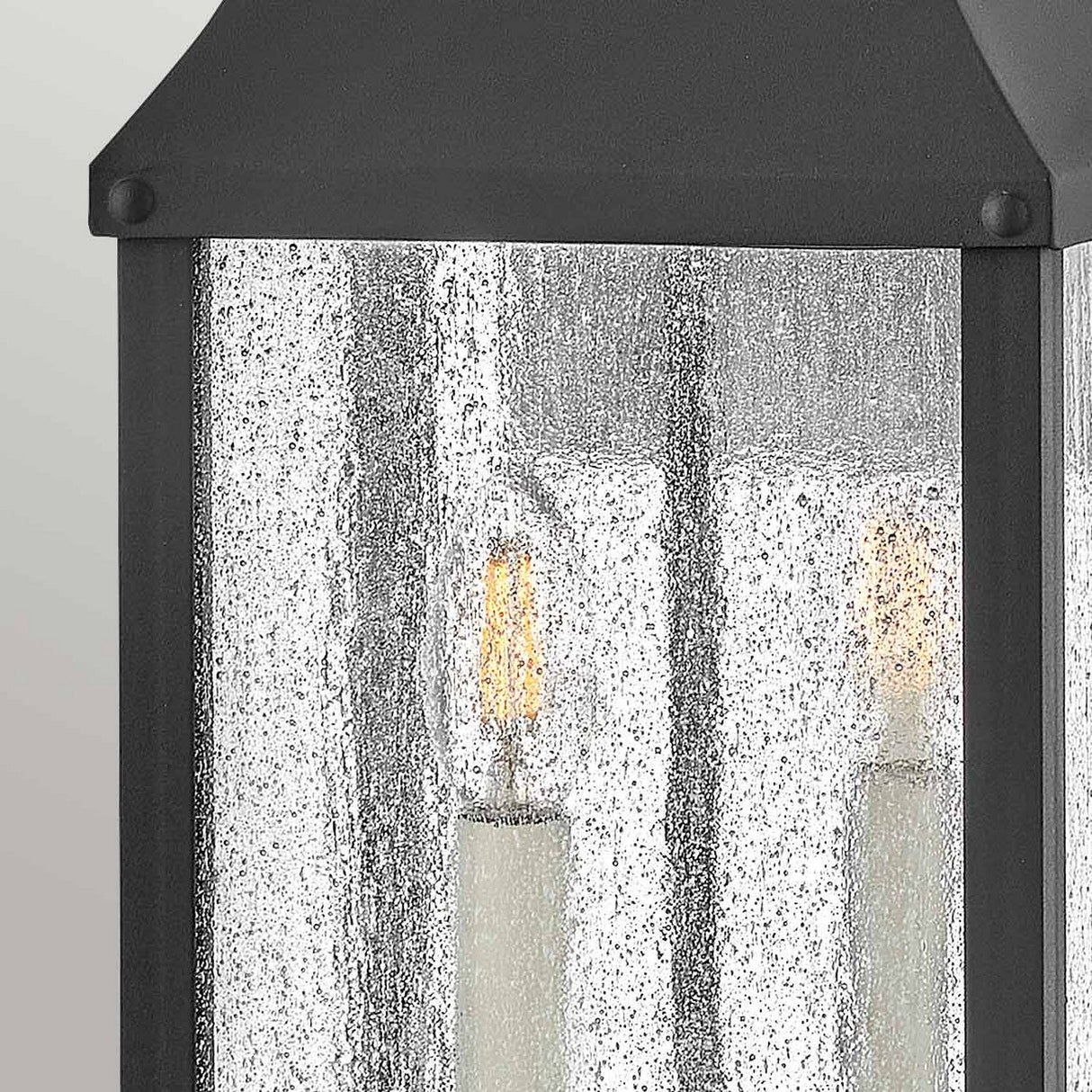 A close-up of the Beacon Hill 2 Light Outdoor Medium Wall Lantern in black aluminum showcases its design with glass panels. Inside, two candle-like bulbs provide a warm glow. The slightly textured, frosted glass gently diffuses the light, set against a background that transitions smoothly from light to dark gray.