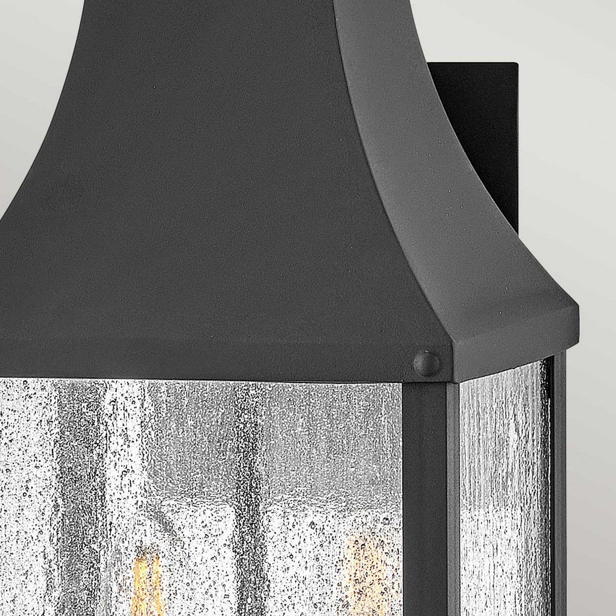 Close-up of a Beacon Hill 2 Light Outdoor Medium Wall Lantern in black aluminum with a textured glass panel, designed in a classic style. The light inside emits a warm glow that highlights the detailed finish of the metal frame, set against a softly blurred background.