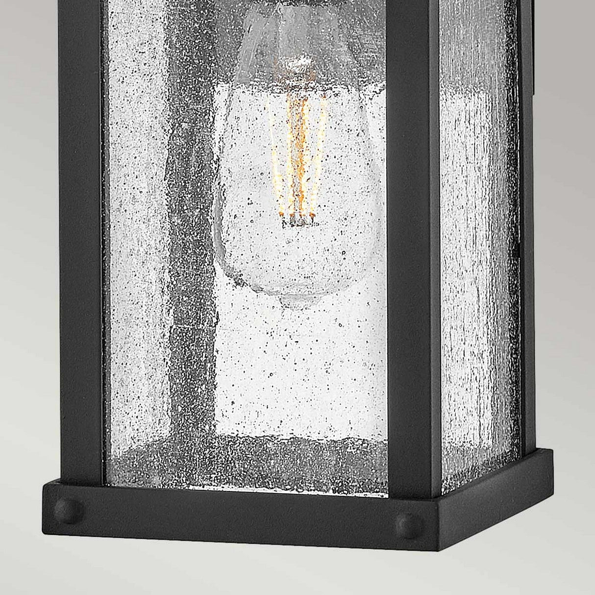 A close-up of the Beacon Hill 1 Light Outdoor Small Wall Lantern in black showcases its clear seeded glass and a vintage-style Edison bulb. The lantern's rectangular design delivers a modern appeal, while the textured glass provides a speckled appearance that enhances its sophisticated charm.