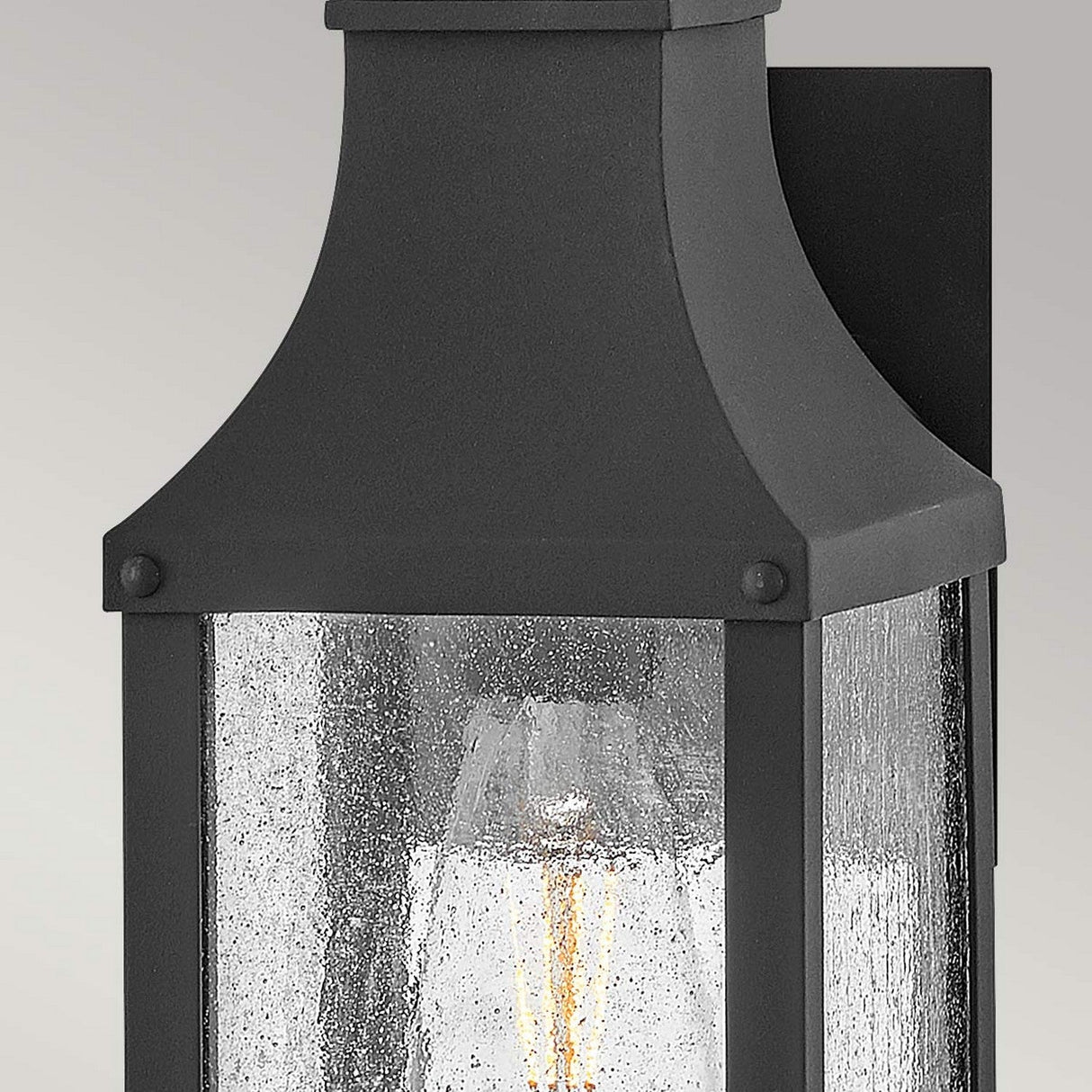 A close-up of the Beacon Hill 1 Light Outdoor Small Wall Lantern in black showcases its seeded glass design. The visible light within casts a warm glow, while its simple and elegant style contrasts beautifully against a neutral background.