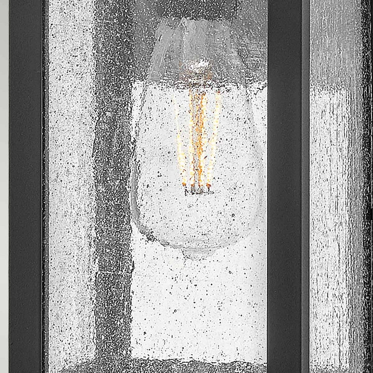 A close-up of the Beacon Hill 1 Light Outdoor Small Wall Lantern in black showcases a vintage-style light bulb with glowing filaments, encased in textured metal. The fixture's museum black hardware and seeded glass emit a warm light, enhancing its antique aesthetic.