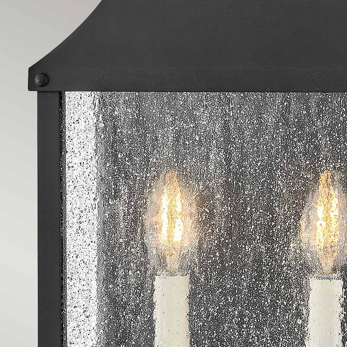 Close-up of the Beacon Hill 2 Light Outdoor Half Wall Lantern - Black, featuring two candle-like bulbs glowing behind a textured, speckled glass panel. The frame is crafted from Museum Black Aluminum, adding a sophisticated touch.
