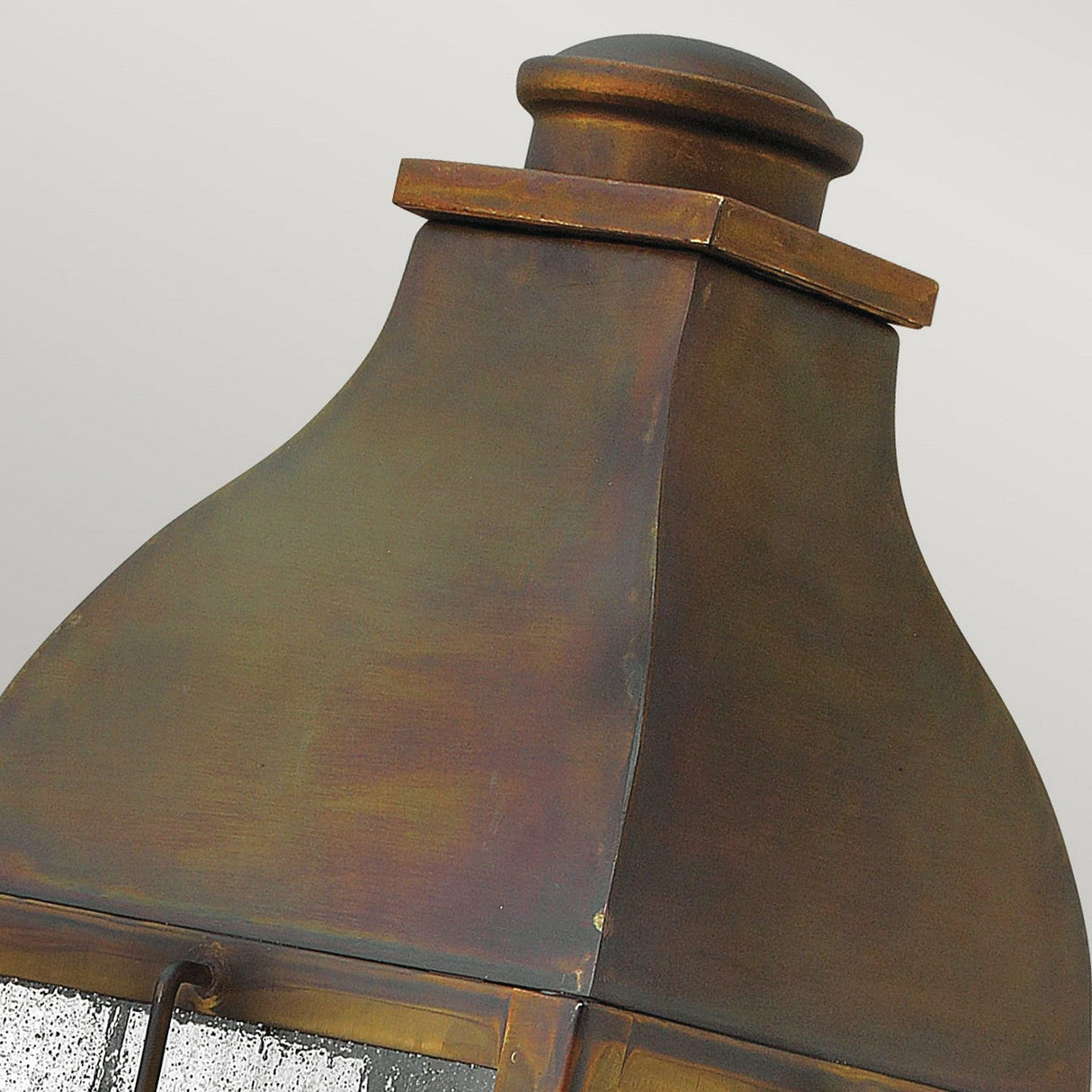 A close-up of the top portion of the Bingham 3 Light Outdoor Large Wall Lantern reveals its antique-style design, boasting a weathered metal construction with a rustic brown finish. The lantern evokes a vintage appeal with its curved structure and slightly domed cap, featuring textured glass panels subtly visible against a plain, light gray backdrop.