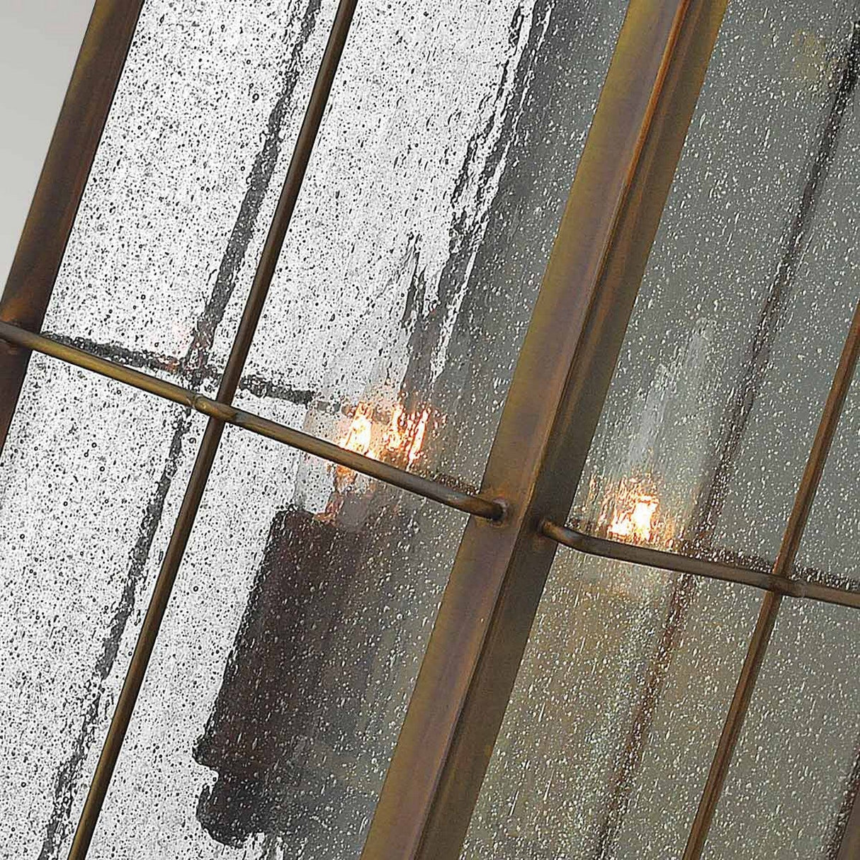 A close-up of a window with raindrops on the glass reveals the Bingham 3 Light Outdoor Large Wall Lanterns just beyond the wet pane. These lights create a warm glow through the textured surface. The window frame appears to be metal, adding an industrial touch with a hint of vintage aesthetic.
