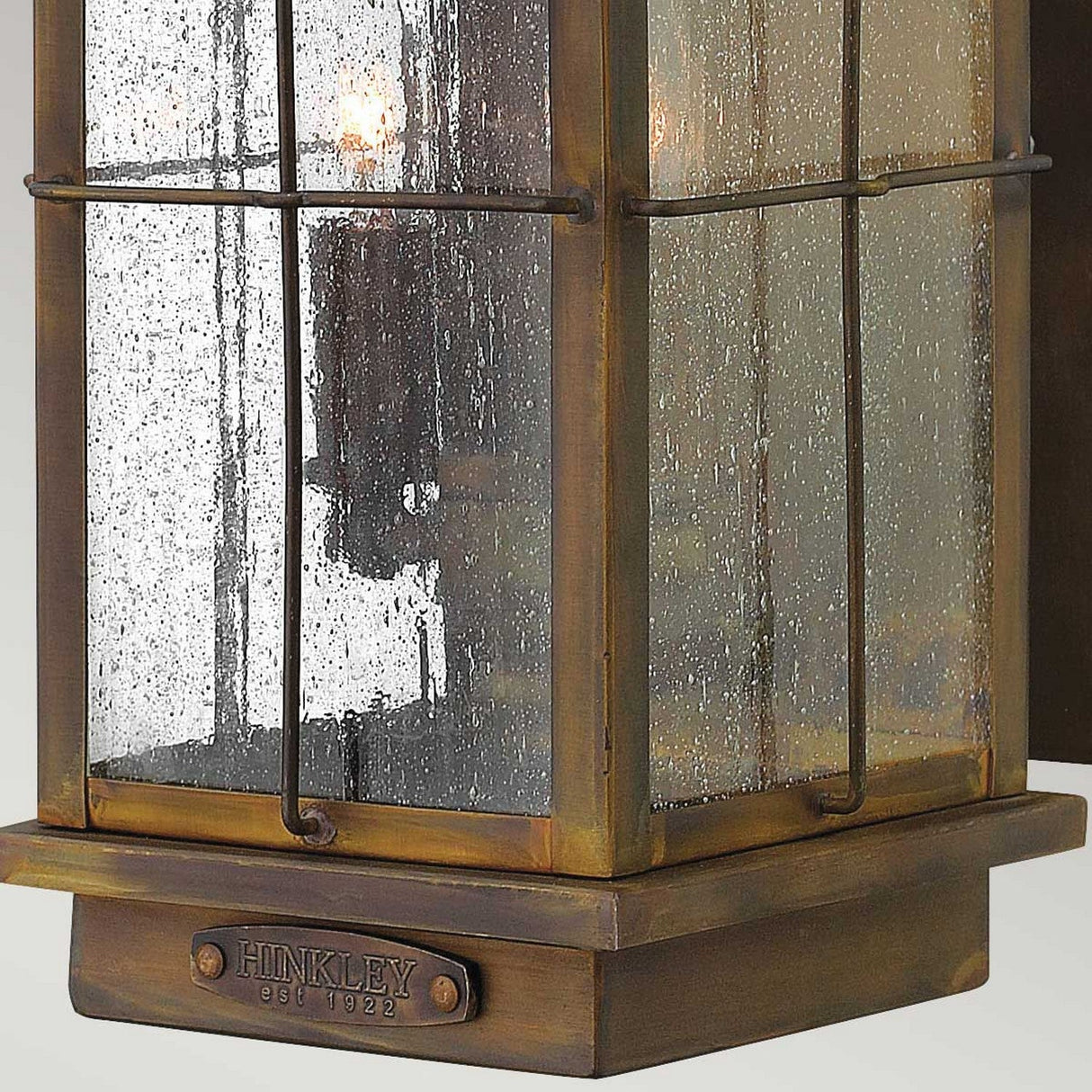 Close-up of the Bingham 3 Light Outdoor Medium Wall Lantern, showcasing a nautical-inspired vintage design with a weathered brass finish and clear seeded glass. The solid brass lighting base has "HINKLEY est. 1922" inscribed on it.