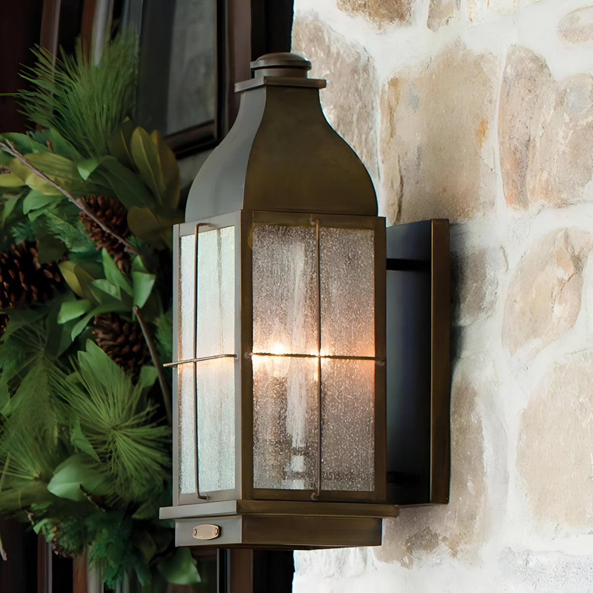 A Bingham 3 Light Outdoor Medium Wall Lantern in brown, featuring a solid brass lighting fixture and glass panels, is mounted on a stone wall. The lantern casts a warm glow as it lights up the area. Greenery accented with pine cones decorates the space beside it, enhancing its vintage charm.